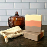 Two handmade layered soap bars in brown, orange, and cream are displayed on a dark wooden table next to a cream mesh soap scrubber and an amber glass pumpkin container, with a white subway tile wall in the background.