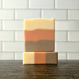 Two handmade layered soap bars stacked on a dark wooden surface, with a white subway tile wall in the background. The soaps feature horizontal stripes of cream, terracotta orange, and brown