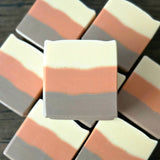 Several soap bars arranged on a dark wooden surface, with additional similar bars blurred in the background. The center bar is in sharp focus and shows horizontal bands of off-white, muted peach, and brown.