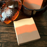 A rectangular bar of handmade soap with three horizontal color bands—cream, orange, and brown—resting on a dark wooden surface next to amber glass pumpkin shaped jar.