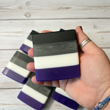 Hand holding a striped soap bar with black, white, and purple layers on a wooden surface.