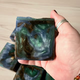 Hand holding a green, blue, and dark brown soap bar with a wooden background