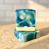 stack of soaps that look like the beach with an oatmeal base and the waves are a swirl of blue, teal, and white. Sitting on a marble counter top with sunlight shining on the soaps.