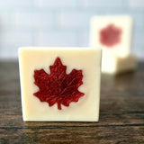Square soap bar sitting up on a dark wooden surface, with a couple of stacked soap bars in the background.  Soap is white with a red maple leaf in the center.