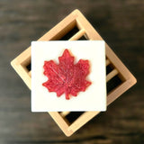 Top down view of a soap bar sitting on a wood slatted soap dish on a dark wood table.  The soap is white with a red maple leaf embedded in the center.