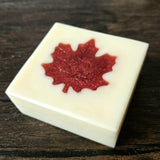 square soap bar sitting on a dark wood table, white shea butter soap bar with a dark red sparkly maple leaf nestled in the center top of the bar.  edges are beveled.