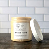 A jar of Pretty Soap Co. French Roast Foaming Sugar Scrub with its white lid leaning against it, set on a dark wooden surface with a white subway tile background.