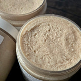 Close-up of a clear plastic jar filled with sugar scrub showcasing the texture, with another similar jar partially visible in the background on a dark wooden surface.