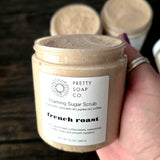 A hand holding a jar of Pretty Soap Co. French Roast Foaming Sugar Scrub with the lid off showing the texture of the scrub, with two more jars blurred in the background on a dark wooden surface.