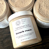A jar of Pretty Soap Co. French Roast Foaming Sugar Scrub on a dark wooden surface lying on its side, surrounded by other jars of the same product without the lids showing the scrub inside.
