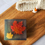 Square soapbar resting on a wooden tray with a woven soap bag next to the soap. Soap has two maple leaves embeded inside a clear soap base, sitting on top of a brown and green soap base.  Gold mica/glitter swims inside the clear soap base.