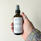 Hand holding a bottle of Lavender Essential Water spray against a light colored wall.