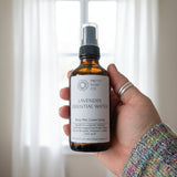 Hand holding a bottle of Lavender Essential Water against a bright window with sheers