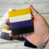 Hand holding a nonbinary flag striped soap bar with a wooden background