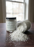 Two jars of bath salts on a wooden surface with some spilled out.