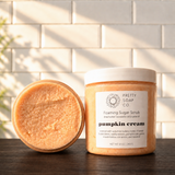 A jar of Pretty Soap Co. Pumpkin Cream Foaming Sugar Scrub stands next to its open lid, showing the textured orange scrub inside.