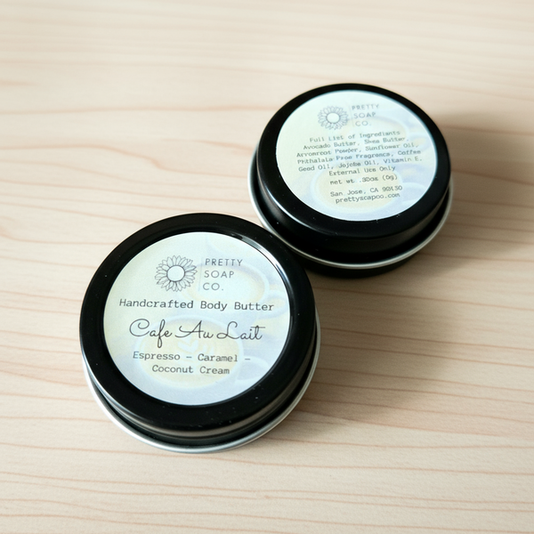 Two mini sampler tins of handcrafted body butter, labeled 'Cafe Au Lait', with black lids on a wooden surface.