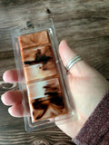 Hand holding a package of brown and black swirled wax melts on a wooden surface