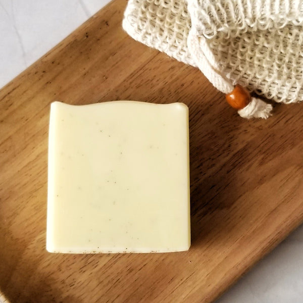 Square soap bar sitting atop a brown wooden tray along side a sisal soap bag.  The soap is yellow with dark brown specks from apricot seed powder. 