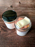 Two jars on a wooden surface, one with a black lid and the other open showing bath salts and reusable tea bag.