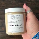 Hand holding one jar of foaming sugar scrub scented in Vanilla Bean, holding the jar over a wooden surface.