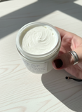 Hand holding a jar of white body butter on a wooden surface